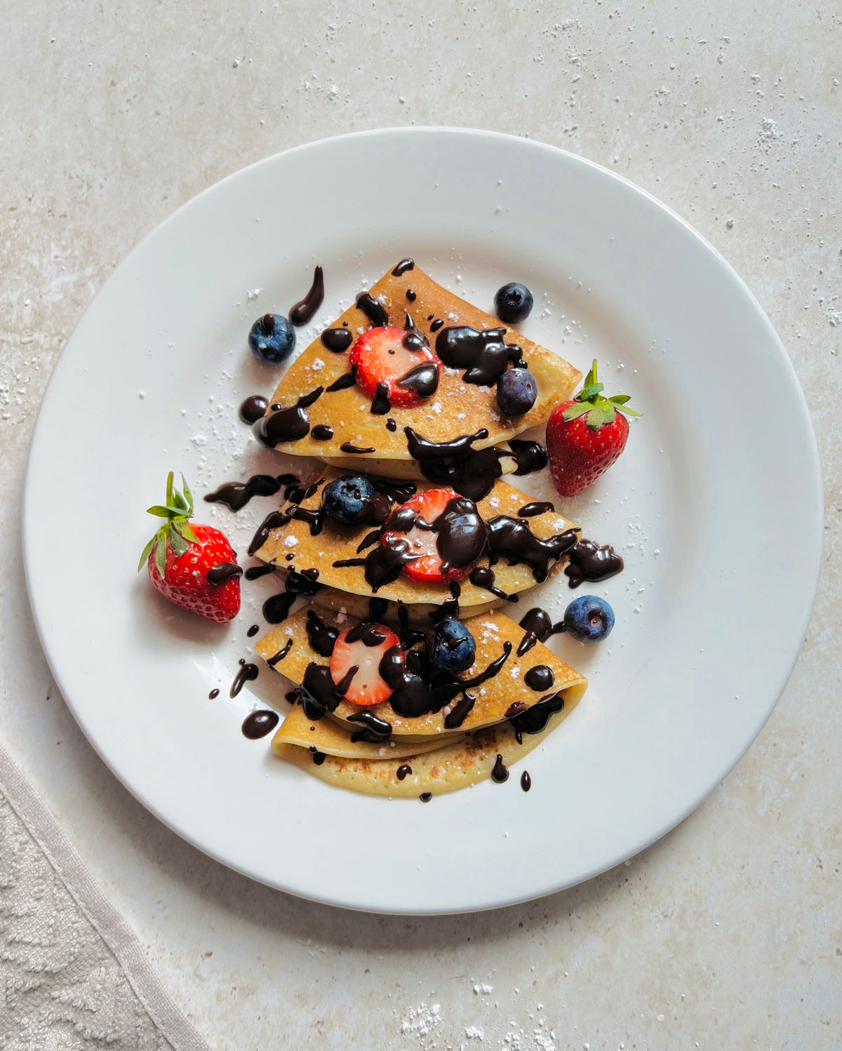 Vegan Crepes on a white plate and served with strawberries, blueberries, icing sugar and dark chocolate sauce